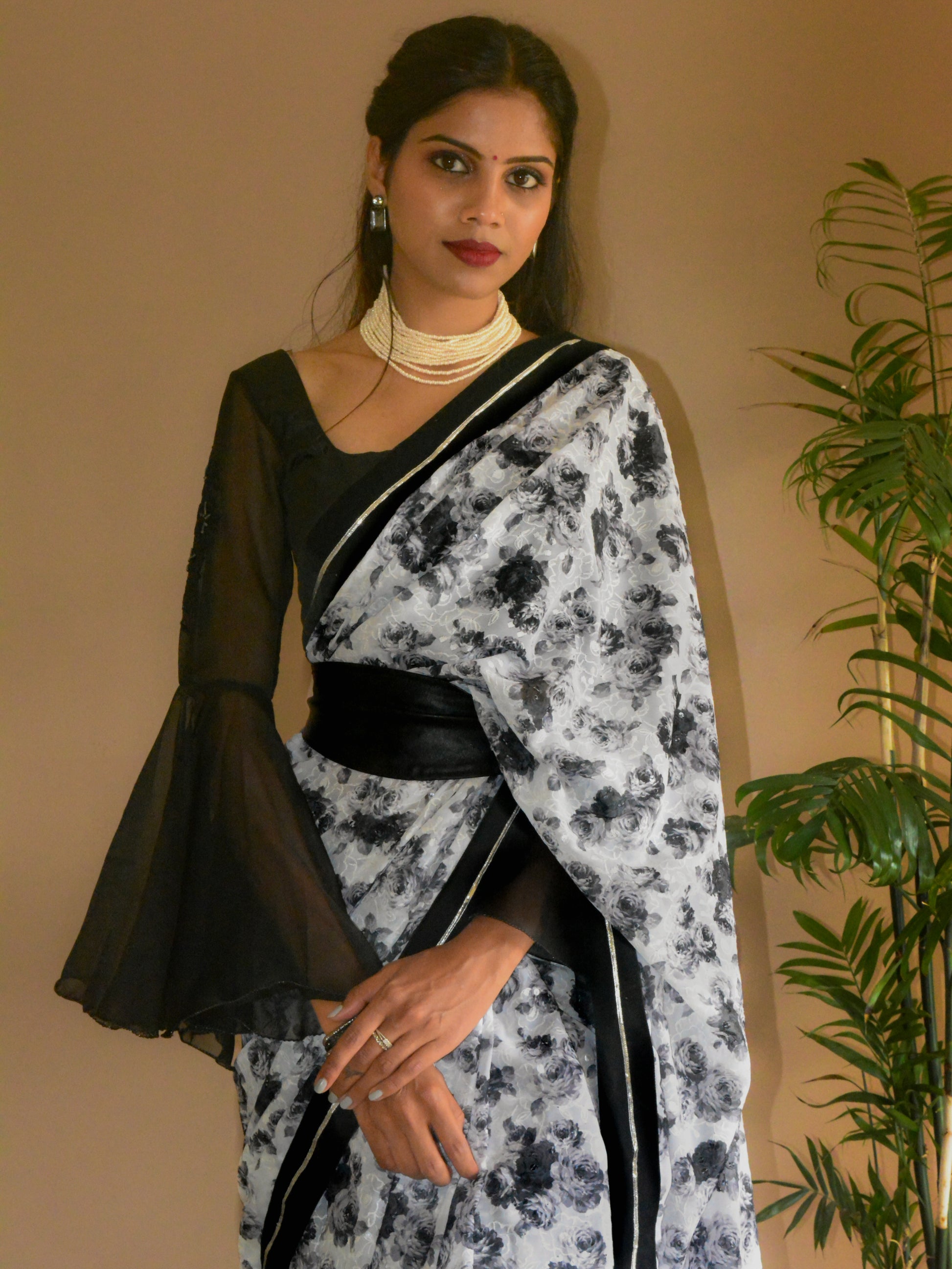 Model wearing a black and white Guldasta Saree featuring a rose print, black velvet border with silver gotta patti, and scattered transparent sequins, evoking a retro 1980s vibe. The saree is paired with black blouse in cotton-silk, flared embroidered sleeves. The Saree is styled with black wide leather belt, and Ratnarani Beaded Choker from the festive collection.