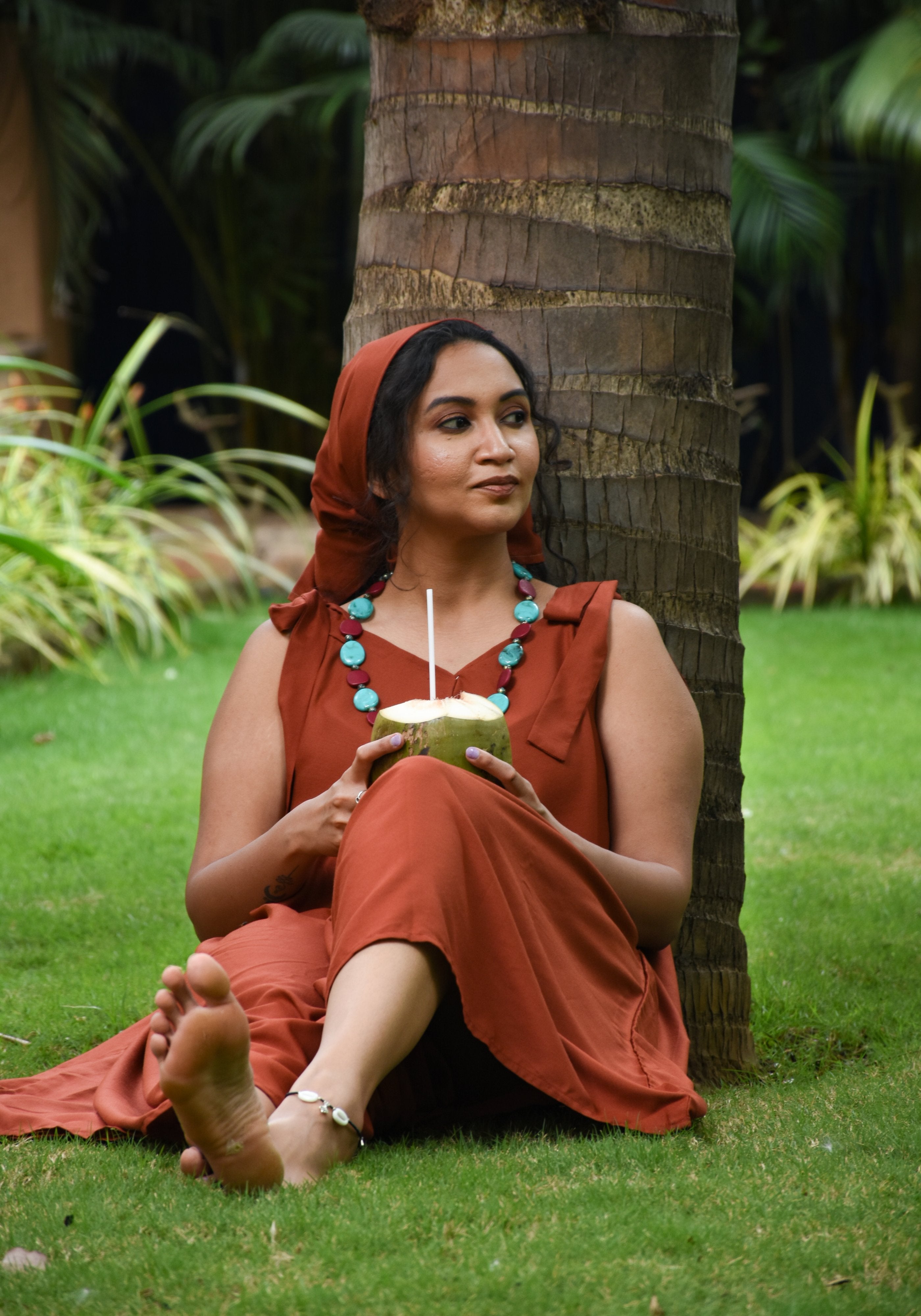 A girl wearing Tie-Shoulder Jumpsuit styled with Opera beaded necklace is sitting on the green grass, under the coconut tree, with tender coconut in her hand, enjoying a quiet moment with herself.