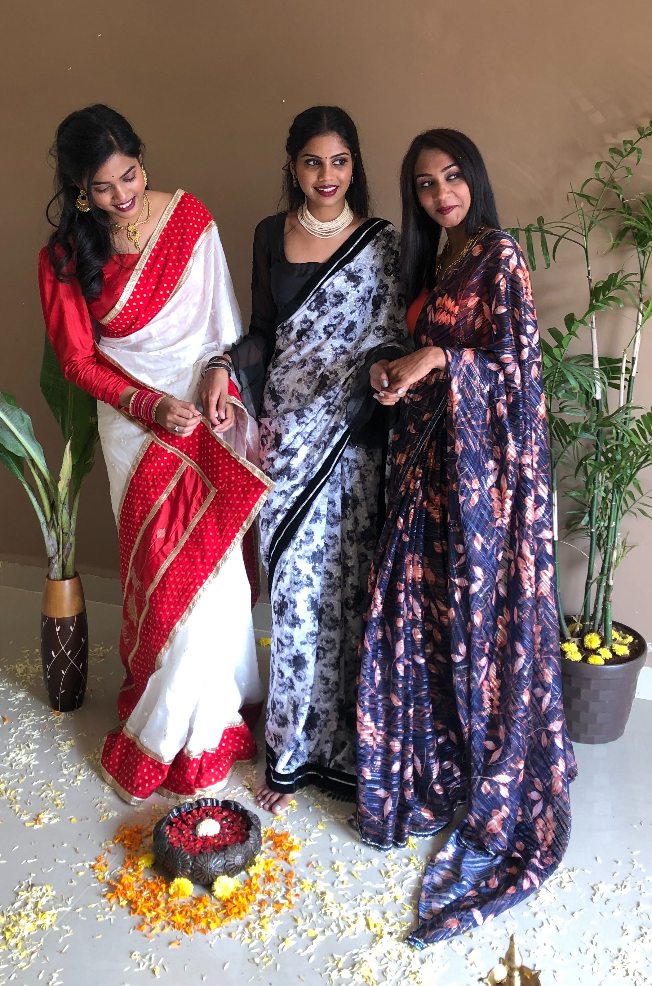 Three women in traditional sarees standing indoors with decorative elements on the floor.