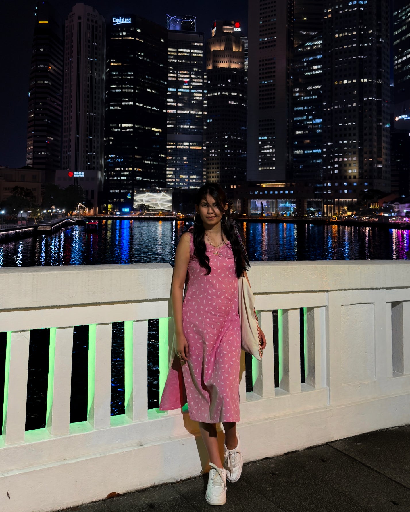 Woman in a pink cotton-linen slip dress standing on a bridge at night with Singapore city skyline in the background