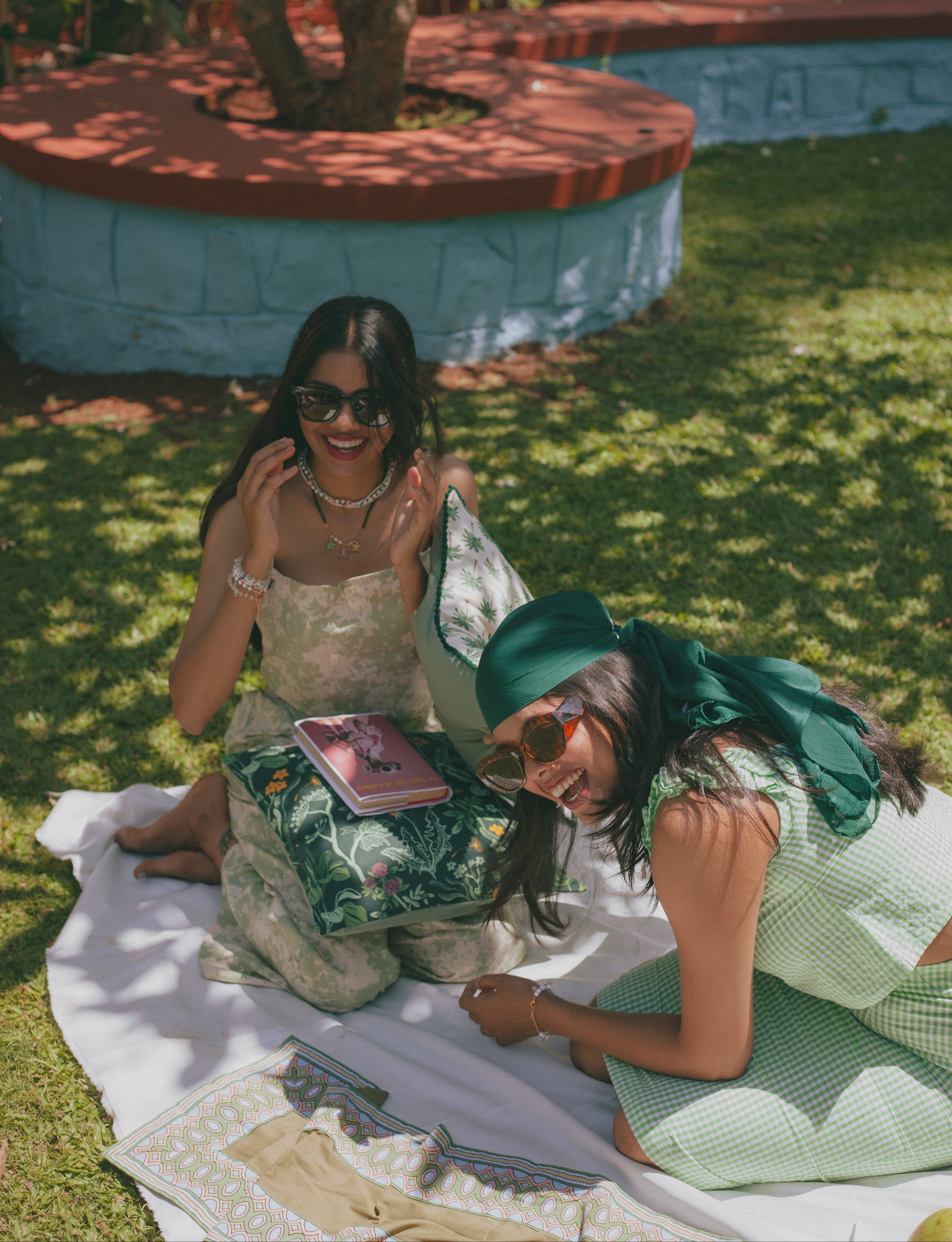 Two women in matching outfits sitting on a blanket outdoors near a pool.