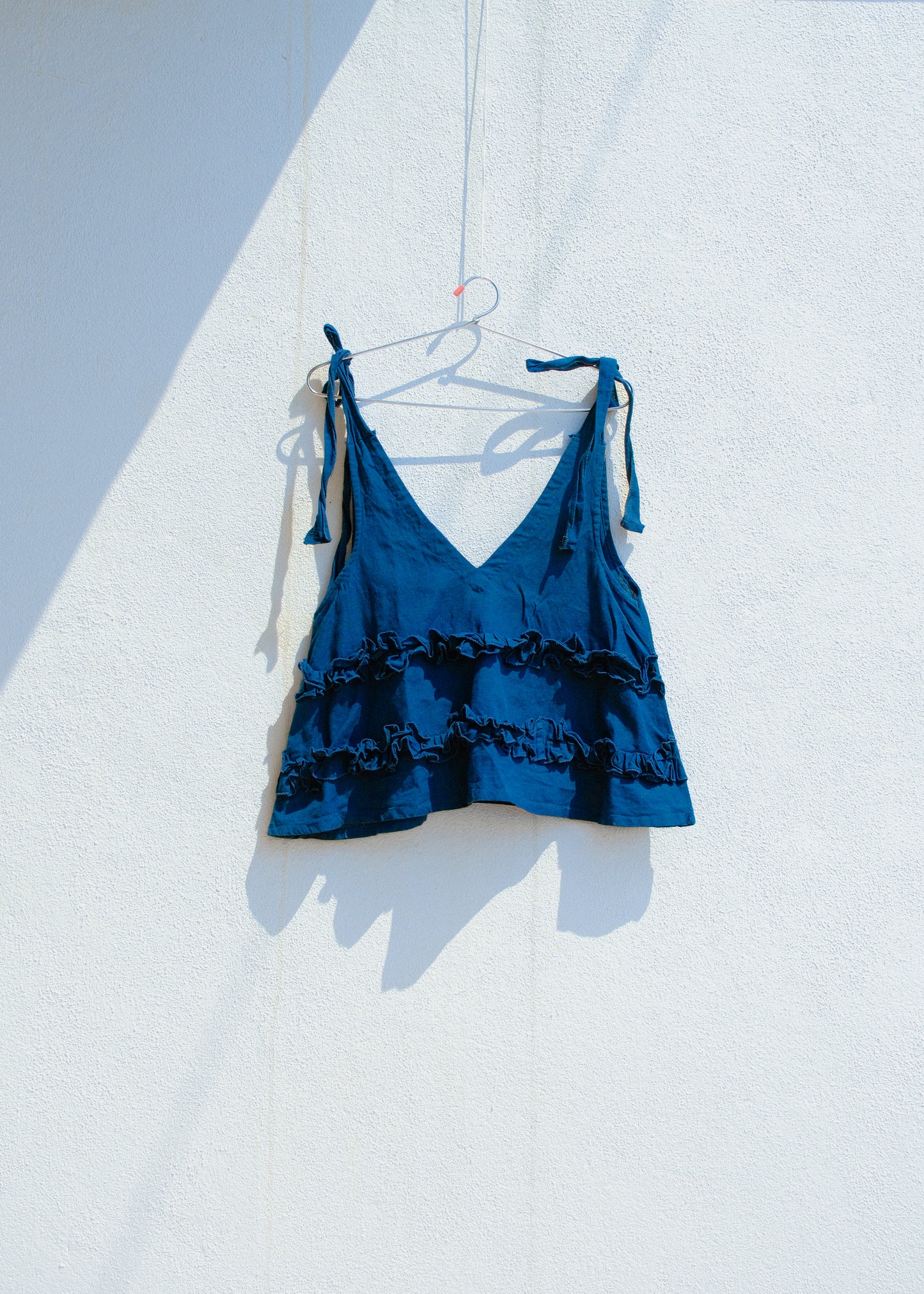 Blue cotton linen tie-shoulder ruffle crop top hanging on hanger against white wall.