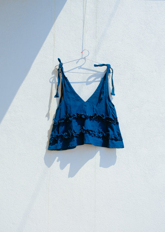 Blue cotton linen tie-shoulder ruffle crop top hanging on hanger against white wall.