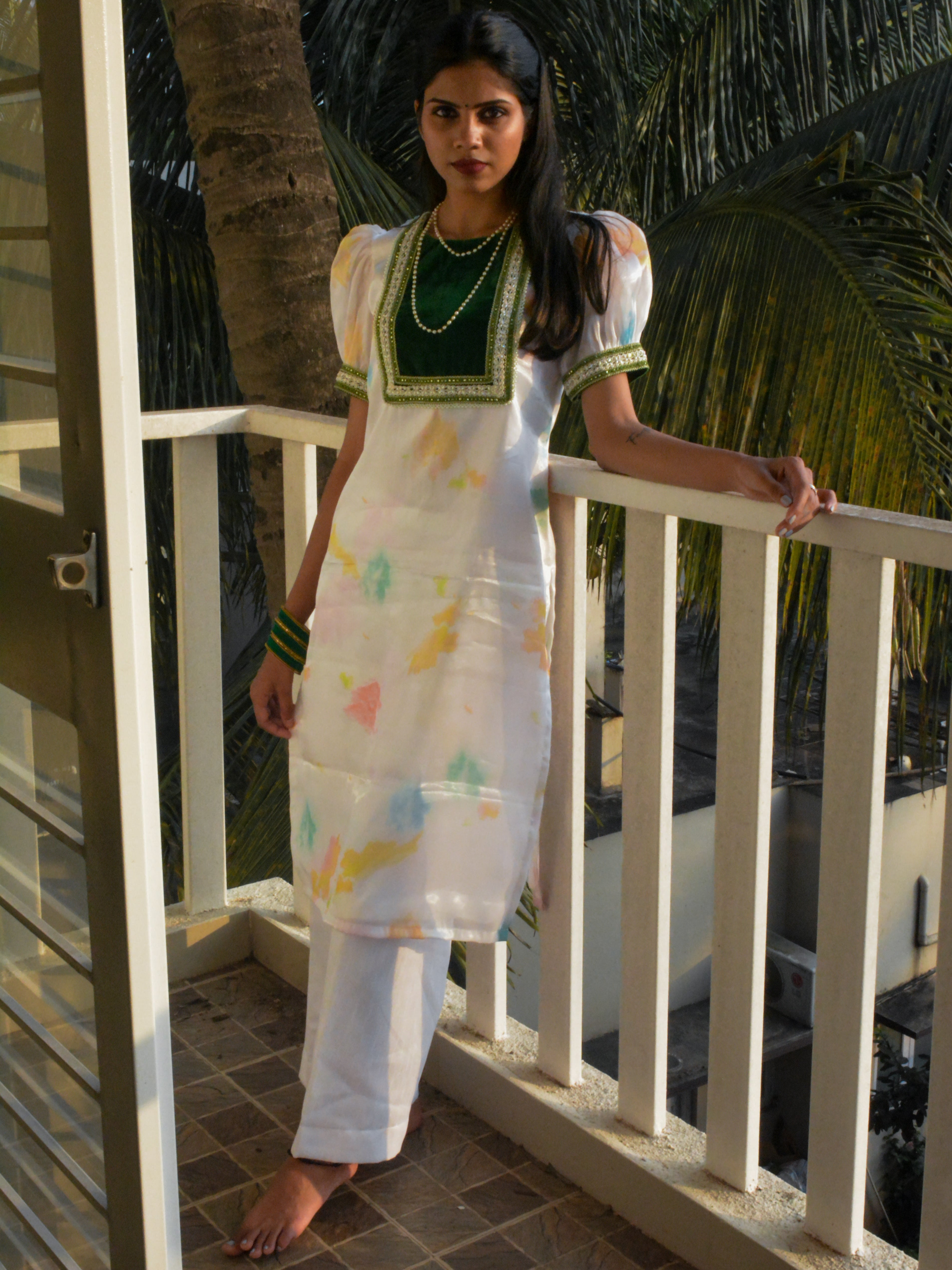 Woman in a white organza kurta paired with white pants standing on a balcony with palm trees in the background