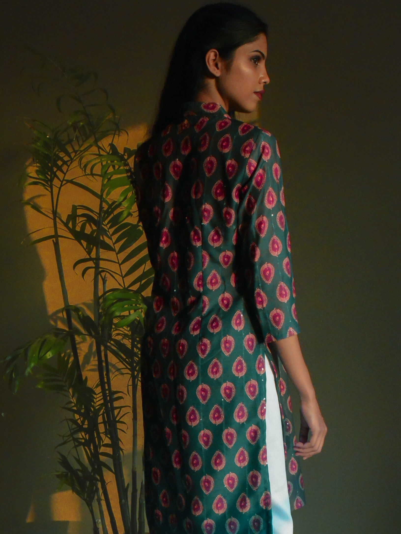 Woman wearing a green printed dress standing against a dark background with a plant to her left.