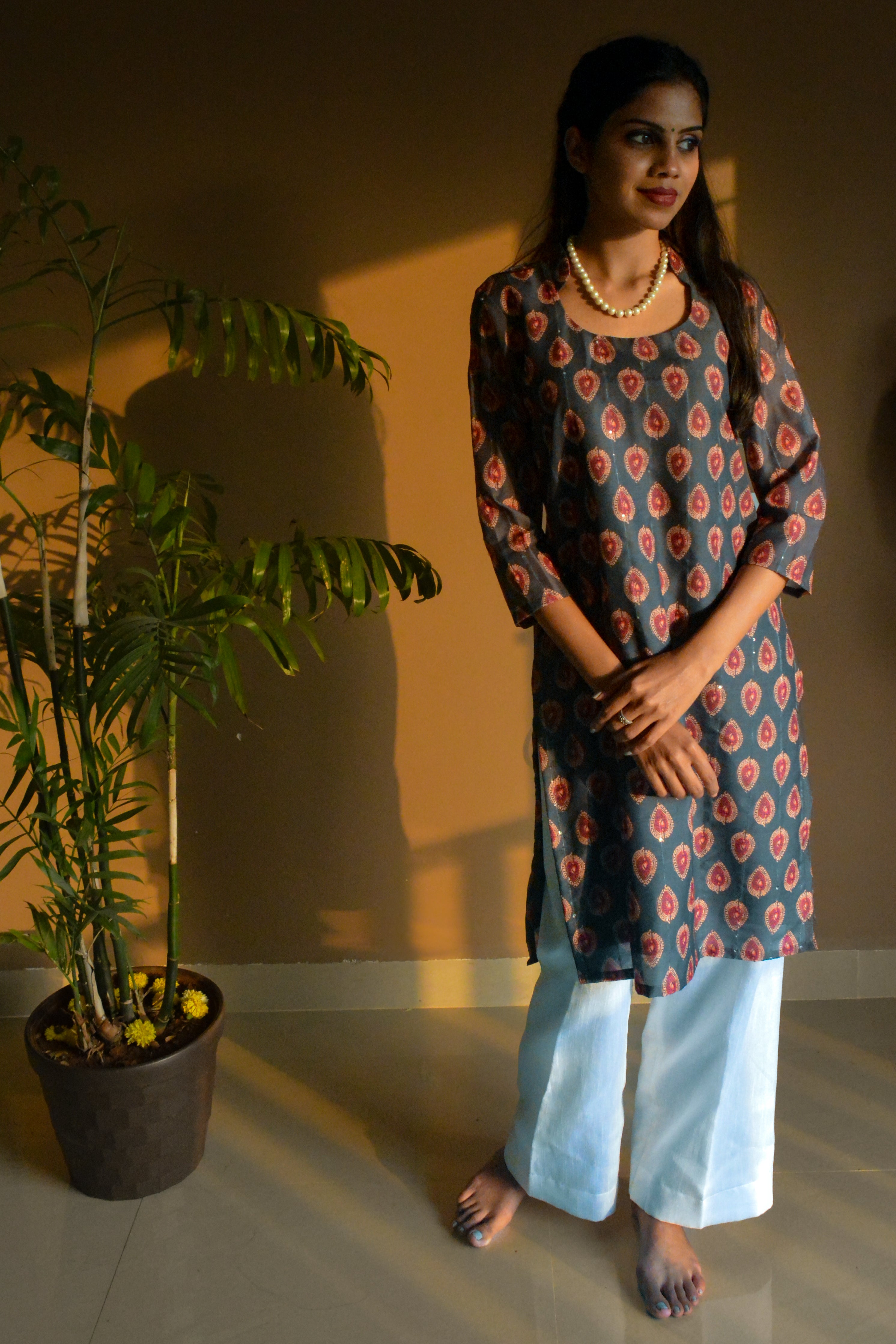 Woman wearing a printed green kurta with white pants standing next to a potted plant indoors.