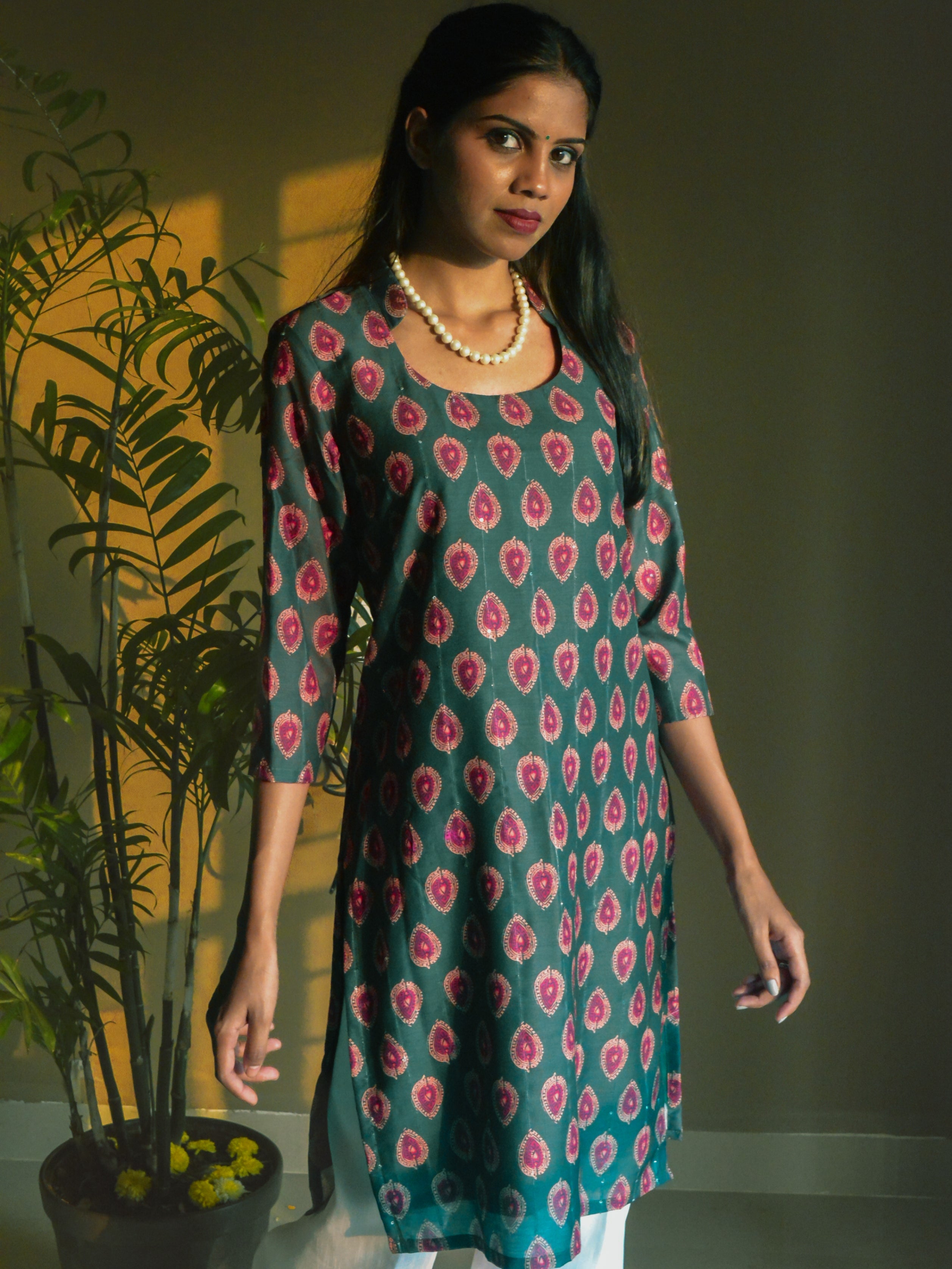 Woman wearing a green and pink printed kurta standing indoors with a plant in the background.