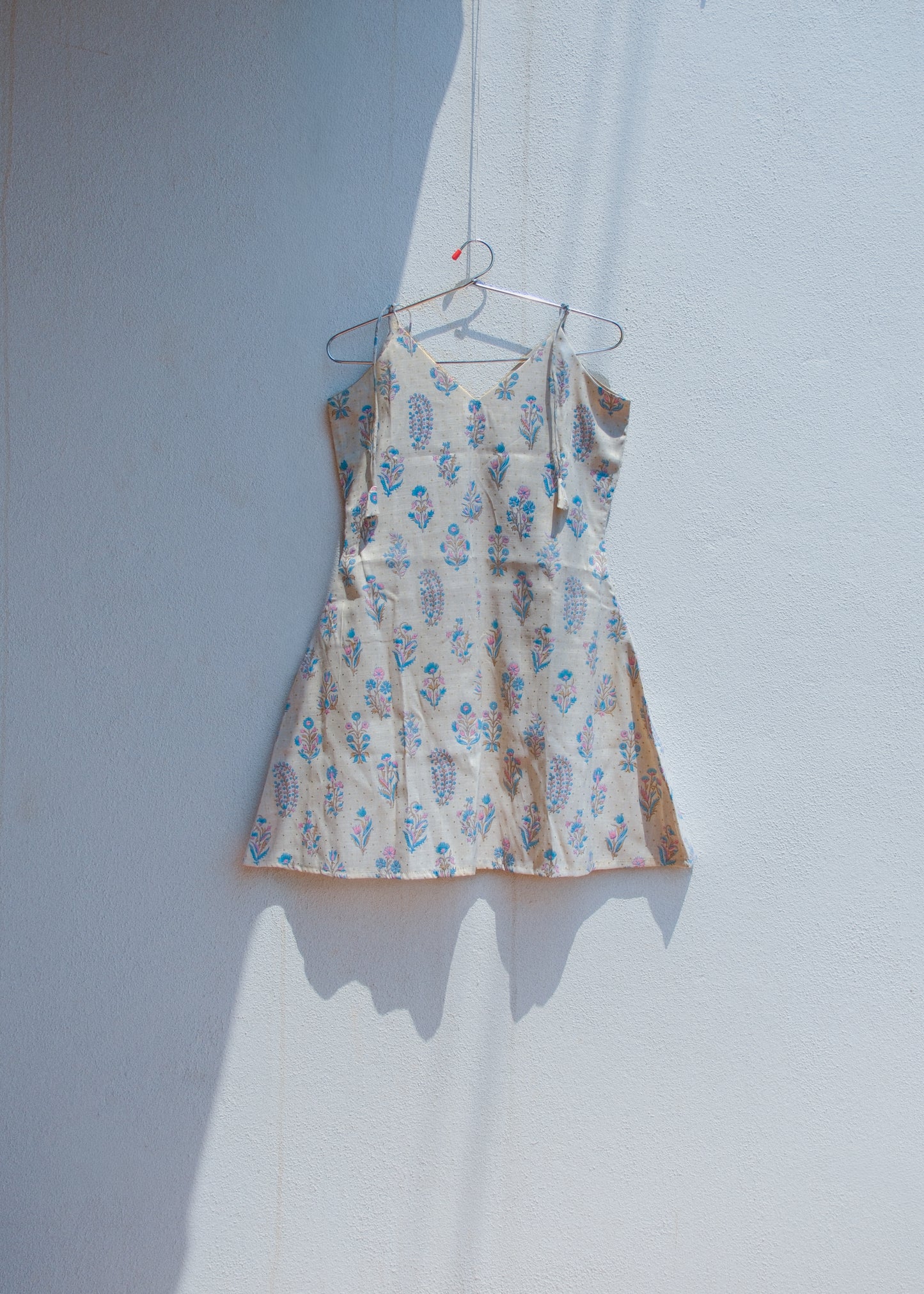 Floral slub cotton A-line dress with tie shoulder straps and V neckline hanging against white wall.