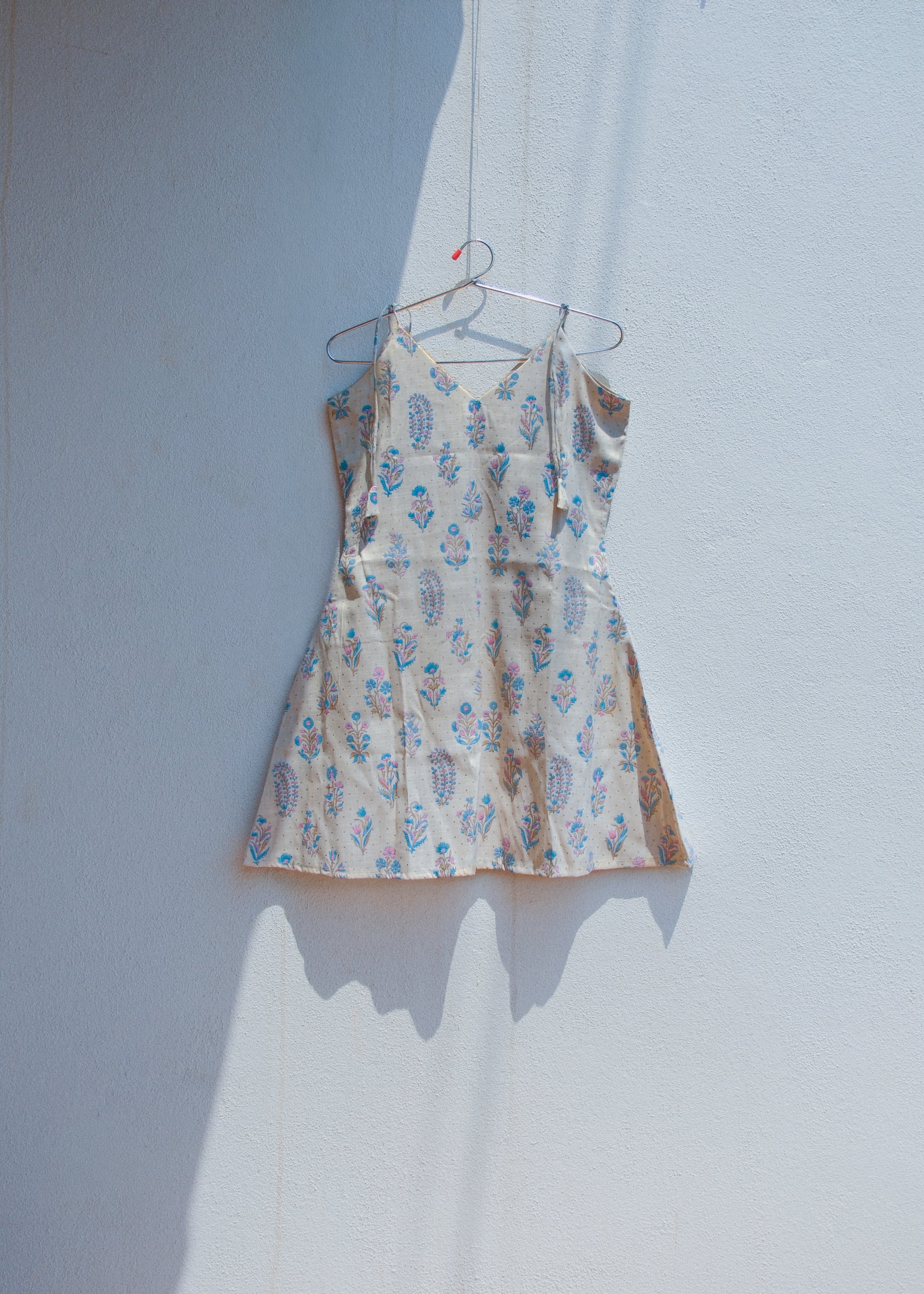 Floral slub cotton A-line dress with tie shoulder straps and V neckline hanging against white wall.