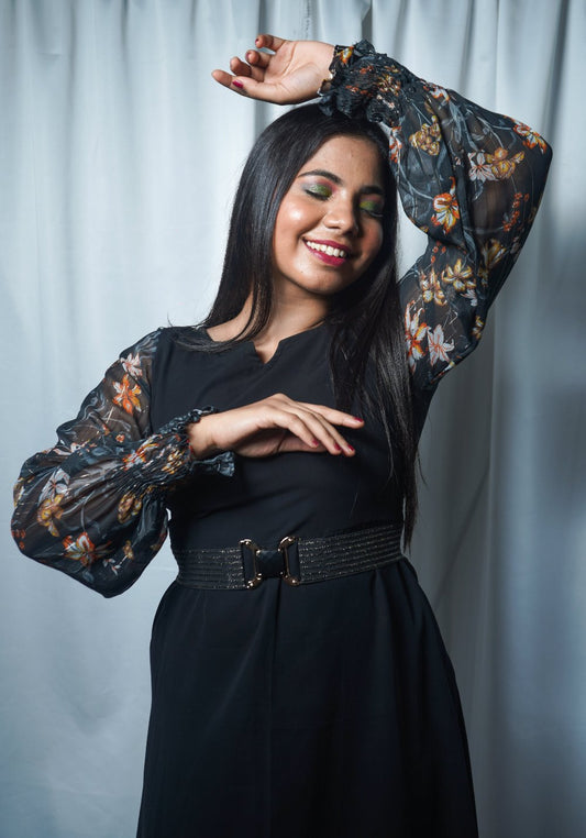 A woman wearing an asymmetrical dress with floral printed long sleeves and a scoop neckline in a charcoal black color.