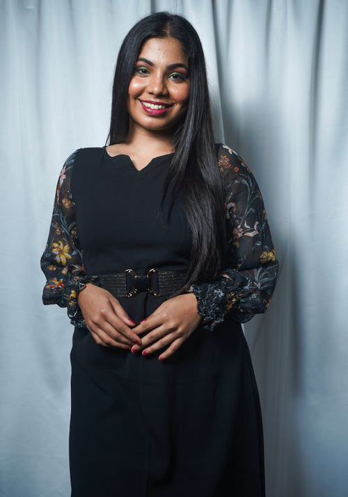A woman wearing an asymmetrical dress with floral printed long sleeves and a scoop neckline in a charcoal black color styled with waist belt.